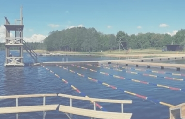 Basen zewnętrzny na jeziorze Väinjärv z torami pływackimi i wysoką wieżą do skoków, wyposażony w pływające pomosty wyprodukowane przez Top Marine, otoczony naturą i błękitnym niebem.